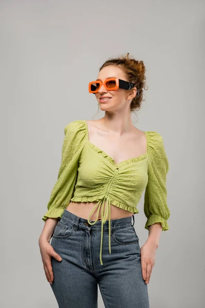 Fröhliche rothaarige Frau in Jeans, grüner Bluse und Sonnenbrille posiert und schaut weg, während sie isoliert auf grauem Hintergrund steht, trendiges Sonnenschutzkonzept, Modemodel — Stockfoto