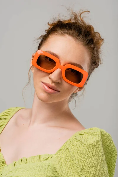 Porträt einer jungen rothaarigen Frau in grüner Bluse und moderner Sonnenbrille, die isoliert vor grauem Hintergrund in die Kamera blickt, trendiges Sonnenschutzkonzept, Modemodel — Stockfoto