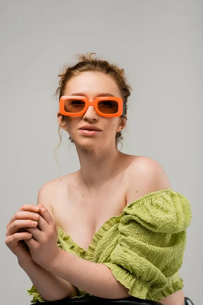 Retrato de mujer de pelo rojo joven con estilo en gafas de sol y blusa verde con hombros desnudos posando mientras está de pie aislado sobre fondo gris, concepto de protección solar de moda, modelo de moda - foto de stock
