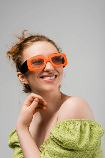 Portrait de jeune femme rousse joyeuse en lunettes de soleil modernes et chemisier vert aux épaules nues regardant loin et posant isolé sur fond gris, concept de protection solaire tendance, modèle de mode — Photo de stock