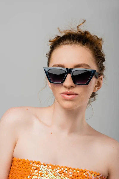 Retrato de mujer de moda y pelirroja en gafas de sol y top naranja con lentejuelas y hombros desnudos de pie aislados sobre fondo gris, concepto de protección solar de moda, modelo de moda - foto de stock