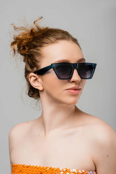Portrait of young red haired and freckled woman in sunglasses and top with sequins looking away while standing isolated on grey background, trendy sun protection concept, fashion model — Stock Photo