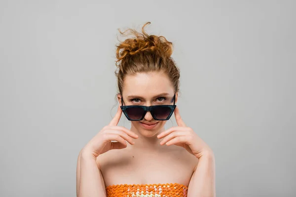 Portrait of stylish young red haired woman with natural makeup in top with sequins and sunglasses looking at camera isolated on grey background, trendy sun protection concept, fashion model — Stock Photo