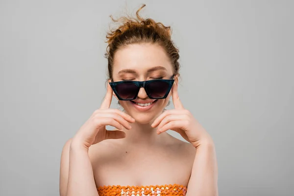 Joven mujer pelirroja alegre con maquillaje natural en gafas de sol y top naranja con lentejuelas cerrando los ojos mientras está de pie aislado sobre fondo gris, concepto de protección solar de moda, modelo de moda - foto de stock
