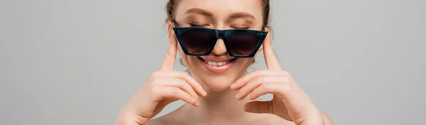 Portrait of young and smiling woman with natural makeup and naked shoulders touching sunglasses while standing isolated on grey background, trendy sun protection concept, fashion model, banner — Stock Photo