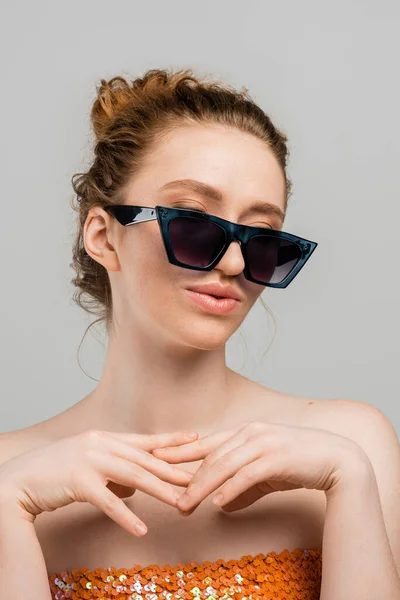 Portrait of young redhead woman with freckles wearing top with sequins and stylish sunglasses while standing isolated on grey background, trendy sun protection concept, fashion model — Stock Photo