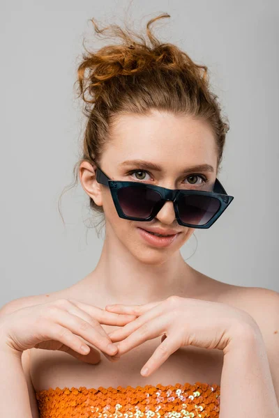 Retrato de joven modelo de pelo rojo con maquillaje natural y hombros desnudos mirando a la cámara y posando en gafas de sol aisladas sobre fondo gris, concepto de protección solar de moda, modelo de moda - foto de stock