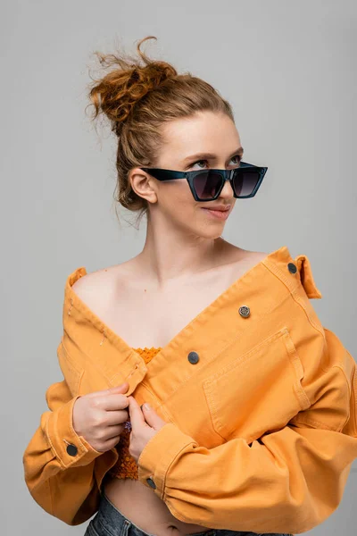 Sorrindo e mulher ruiva jovem na moda em óculos de sol e casaco de ganga laranja olhando para longe enquanto posando isolado em fundo cinza, conceito de proteção solar na moda, modelo de moda — Fotografia de Stock