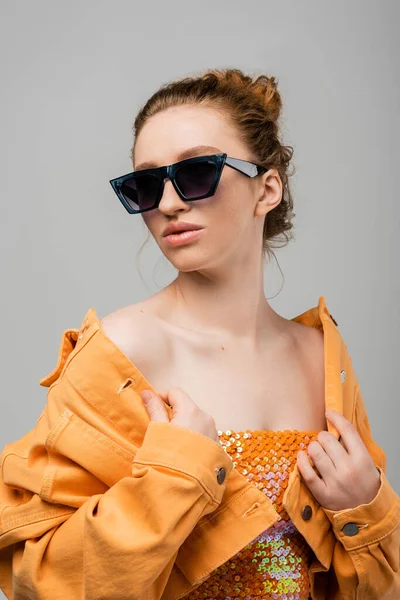 Fashionable redhead woman with natural makeup in sunglasses and top with sequins touching orange jacket while standing isolated on grey background, trendy sun protection concept, fashion model — Stock Photo