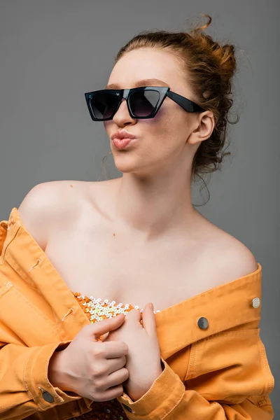 Portrait de jeune femme rousse en lunettes de soleil, top à paillettes et veste en denim orange boudant les lèvres et posant isolée sur fond gris, concept de protection solaire tendance, mannequin — Photo de stock