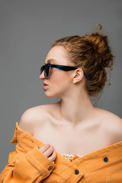 Seitenansicht einer jungen rothaarigen Frau in Sonnenbrille und orangefarbener Jeansjacke mit nackten Schultern, die isoliert auf grauem Hintergrund wegschaut, trendiges Sonnenschutzkonzept, Modemodel — Stockfoto