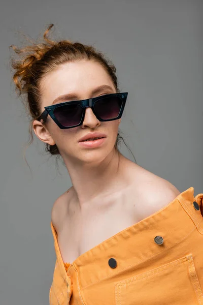 Porträt eines rothaarigen und sommersprossigen Models in Sonnenbrille und orangefarbener Jeansjacke, das isoliert auf grauem Hintergrund posiert und steht, trendiges Sonnenschutzkonzept, Modemodel — Stockfoto