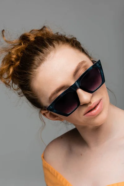Retrato de mujer joven y pelirroja de moda con maquillaje natural usando gafas de sol mientras está de pie aislado sobre fondo gris, concepto de protección solar de moda, modelo de moda - foto de stock