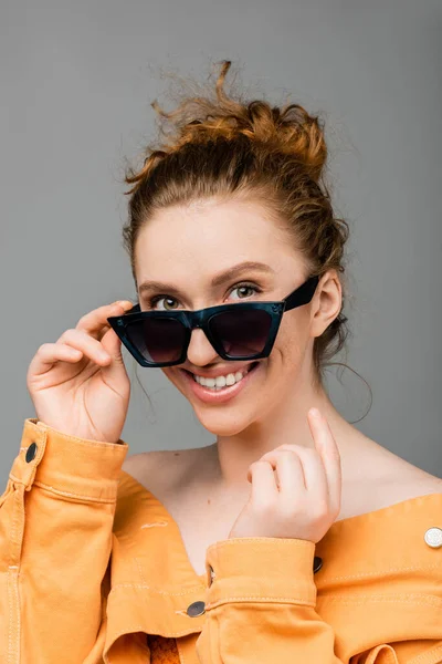 Cheerful young redhead woman with natural makeup touching sunglasses and wearing orange denim jacket while standing isolated on grey background, trendy sun protection concept, fashion model — Stock Photo