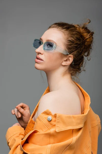 Модная молодая рыжеволосая женщина в солнцезащитных очках и оранжевой джинсовой куртке с голым плечом, отводящим вдаль и стоящая изолированной на сером фоне, триумфальная концепция защиты от солнца, модель — стоковое фото