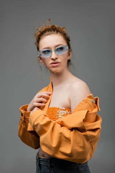 Стильная молодая рыжая женщина в солнцезащитных очках, топ с блестками и оранжевой джинсовой курткой с обнаженным плечом, стоящая изолированно на сером фоне, модная концепция защиты от солнца, модель — стоковое фото