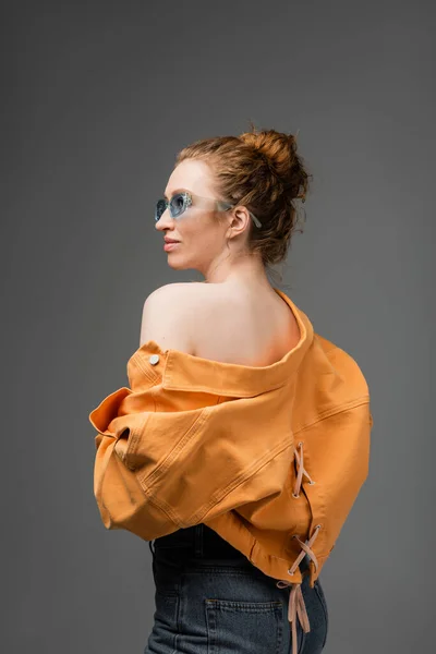 Elegante mujer de pelo rojo joven en gafas de sol y chaqueta de mezclilla naranja con hombro desnudo mirando hacia otro lado mientras está de pie aislado sobre fondo gris, concepto de protección solar de moda, modelo de moda - foto de stock