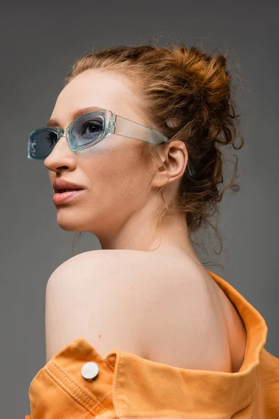 Porträt einer rothaarigen und sommersprossigen jungen Frau in Sonnenbrille und orangefarbener Jeansjacke mit nackter Schulter, die isoliert auf grauem Hintergrund steht, trendiges Sonnenschutzkonzept, Modemodel — Stockfoto