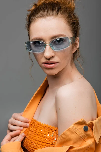 Confiant jeune femme rousse aux lunettes de soleil posant en haut avec des paillettes et une veste en denim orange tout en étant isolé sur fond gris, concept de protection solaire tendance, modèle de mode — Photo de stock