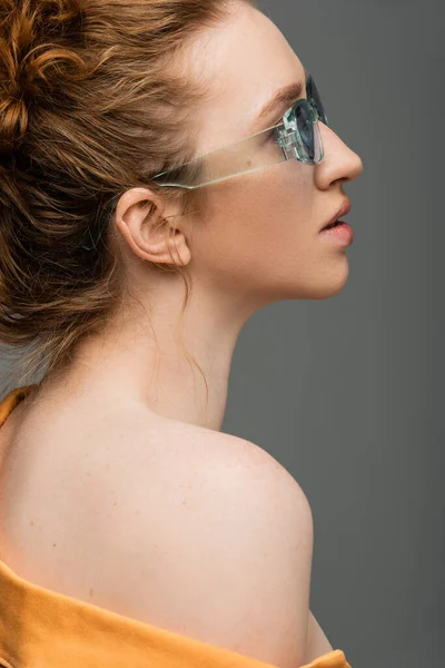 Side view of young red haired and freckled model with natural makeup and naked shoulder posing in sunglasses while standing isolated on grey background, trendy sun protection concept, fashion model — Stock Photo