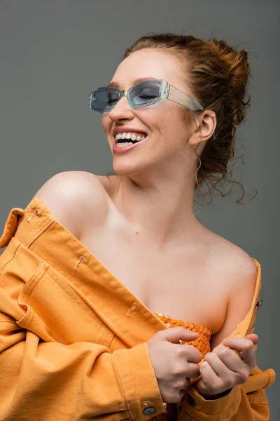 Mulher ruiva alegre e elegante em óculos de sol azuis e casaco de ganga laranja com ombros nus rindo isolado em fundo cinza, conceito de proteção solar na moda — Fotografia de Stock
