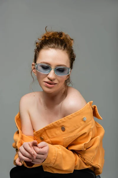 Mujer sonriente y elegante pelirroja en gafas de sol posando en chaqueta de mezclilla naranja con hombros desnudos aislados sobre fondo gris, concepto de protección solar de moda, modelo de moda - foto de stock