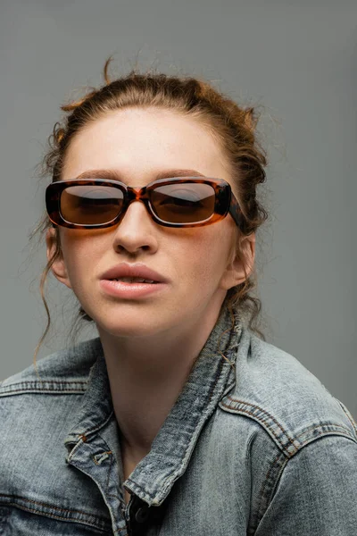Portrait de jeune femme rousse élégante avec des taches de rousseur posant dans des lunettes de soleil et une veste en denim et debout isolé sur fond gris, concept de protection solaire à la mode, modèle de mode — Photo de stock