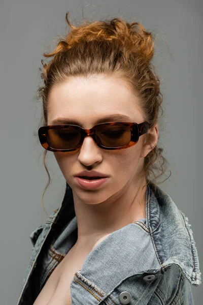 Стильная и уверенная в себе молодая рыжая модель с натуральным макияжем в солнцезащитных очках, позирующая в джинсовой куртке и стоящая изолированная на сером фоне, модная концепция защиты от солнца, модель — стоковое фото