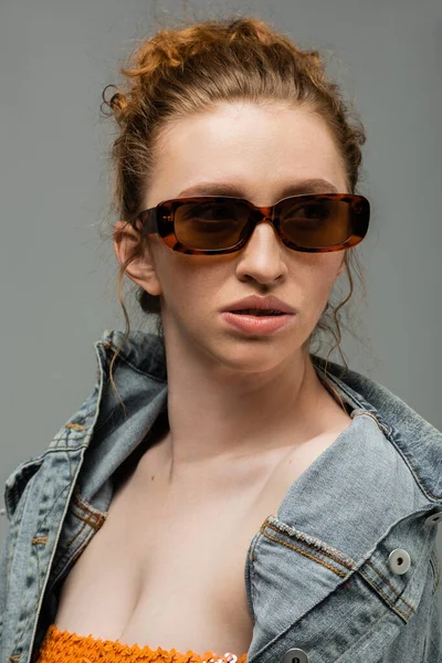 Впевнена молода руда волохата жінка з натуральним макіяжем носить сонцезахисні окуляри та джинсову куртку і дивиться ізольовано на сірому фоні, концепція захисту від сонця, модель моди — стокове фото