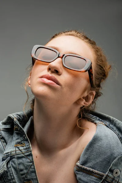 Portrait de jeune femme aux cheveux roux et aux taches de rousseur avec des lunettes de soleil élégantes et une veste en denim debout sous un éclairage isolé sur fond gris, concept de protection solaire tendance, modèle de mode — Photo de stock