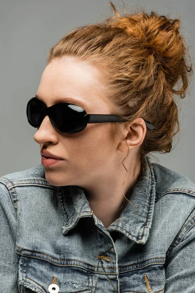 Portrait of stylish freckled and red haired woman in sunglasses and denim jacket posing and standing isolated on grey background, trendy sun protection concept, fashion model — Stock Photo