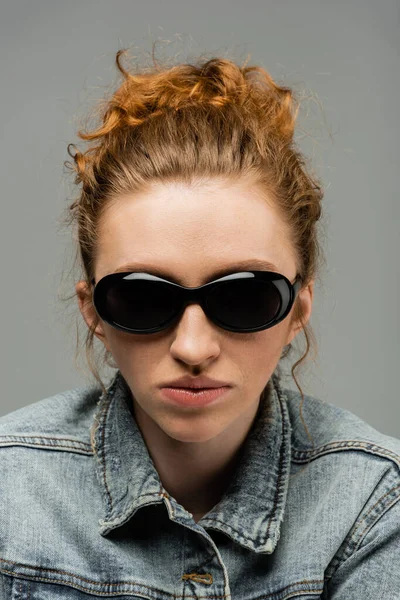 Portrait of young redhead and freckled woman with natural makeup posing in sunglasses and denim jacket while standing isolated on grey background, trendy sun protection concept, fashion model — Stock Photo