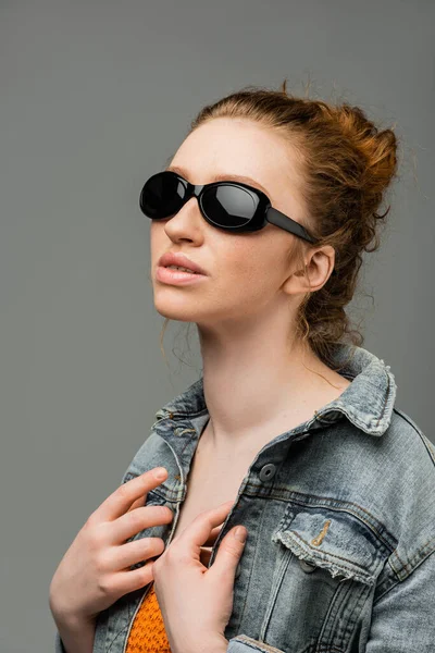Mujer joven pelirroja y pecosa en gafas de sol elegantes posando en chaqueta de mezclilla y parte superior con lentejuelas y de pie aislado sobre fondo gris, concepto de protección solar de moda, modelo de moda - foto de stock