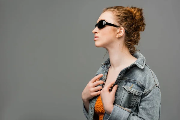 Young redhead model with natural makeup posing in sunglasses and top with sequins while touching denim jacket isolated on grey background, trendy sun protection concept, fashion model — Stock Photo