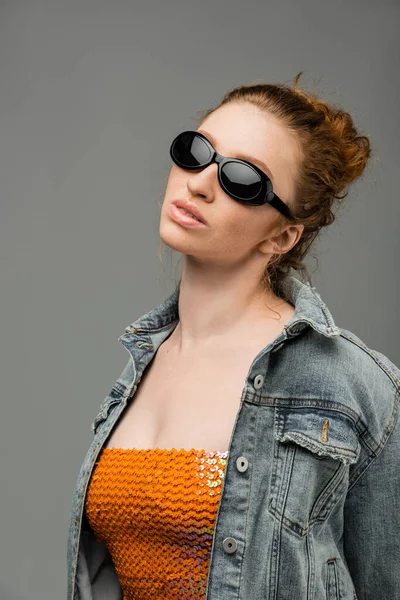 Mujer pelirroja joven confiada y elegante en gafas de sol, parte superior con lentejuelas y chaqueta de mezclilla de pie y posando aislado sobre fondo gris, concepto de protección solar de moda, modelo de moda - foto de stock
