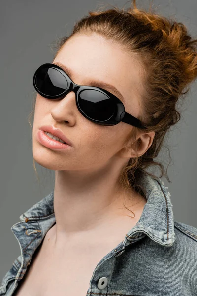 Портрет молодой рыжей и веснушчатой женщины в модных солнцезащитных очках и джинсовой куртке, стоящей изолированно на сером фоне, модная концепция защиты от солнца, модель — стоковое фото