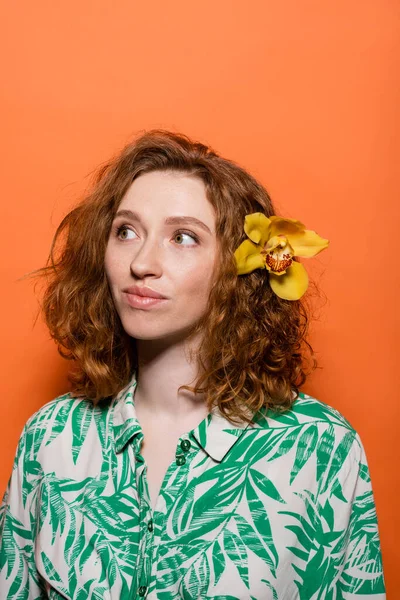 Young redhead and freckled woman with orchid flower in hair wearing blouse with floral print and looking away on orange background, summer casual and fashion concept, Youth Culture — Stock Photo