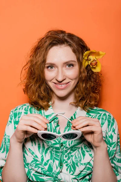 Cheerful young redhead woman with natural makeup and orchid flower in hair looking at camera and holding sunglasses on orange background, summer casual and fashion concept, Youth Culture — Stock Photo