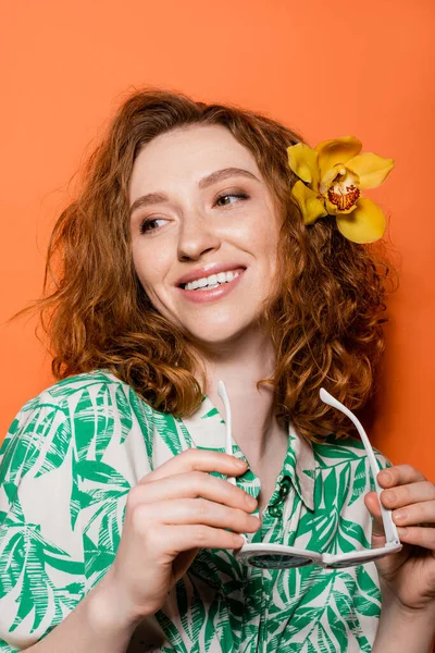 Positive young redhead woman with orchid flower in hair wearing blouse with floral pattern and holding sunglasses on orange background, summer casual and trendy concept, Youth Culture — Stock Photo