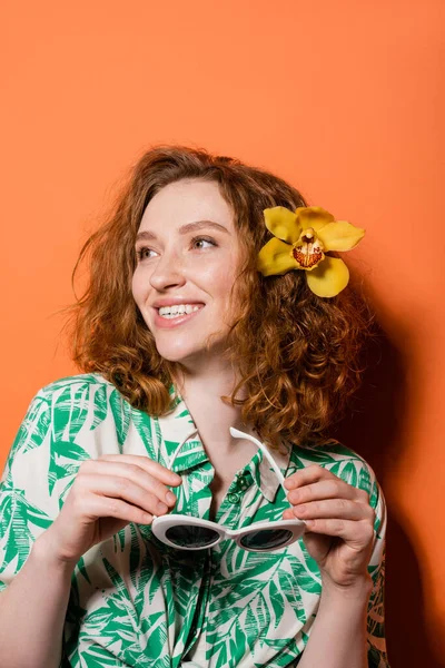 Smiling young redhead woman with orchid flower in hair wearing blouse with floral print and holding sunglasses while standing on orange background, summer casual and fashion concept, Youth Culture — Stock Photo