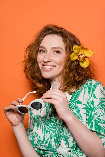 Positive junge rothaarige Frau mit Orchideenblüte im Haar, die in die Kamera schaut und Sonnenbrille hält und auf orangefarbenem Hintergrund steht, Sommer-Freizeit- und Modekonzept, Jugendkultur — Stockfoto