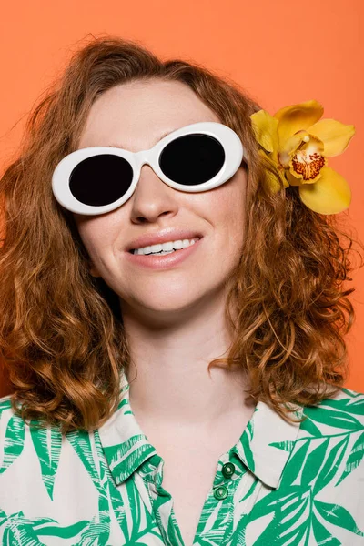 Retrato de mujer joven alegre y pelirroja con flor de orquídea en el pelo con gafas de sol y de pie sobre fondo naranja, verano casual y concepto de moda, Cultura Juvenil - foto de stock