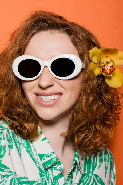 Porträt einer positiven jungen rothaarigen Frau mit Orchideenblume im Haar, die mit Sonnenbrille und Bluse mit floralem Print auf orangefarbenem Hintergrund posiert, sommerliches Freizeit- und Modekonzept, Jugendkultur — Stockfoto