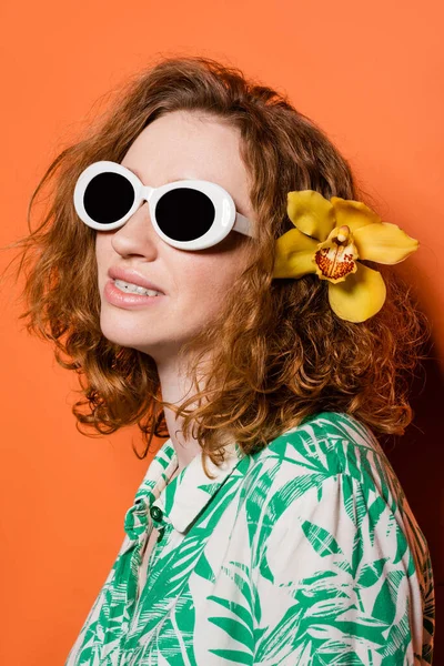 Молодая модная рыжая женщина в солнечных очках, блузка с цветочным шрифтом и цветок орхидеи в волосах улыбается, стоя на оранжевом фоне, летняя повседневная и модная концепция, молодежная культура — стоковое фото
