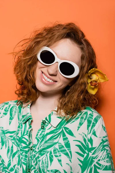 Jeune modèle rousse avec fleur d'orchidée dans les cheveux et les lunettes de soleil et chemisier avec motif floral souriant et debout sur fond orange, concept décontracté et mode d'été, Culture de la jeunesse — Photo de stock