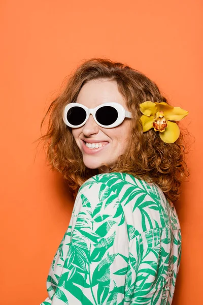 Mujer joven positiva con pelo rojo y flor de orquídea posando en gafas de sol y blusa con estampado floral sobre fondo naranja, concepto casual y de moda de verano, Cultura Juvenil - foto de stock