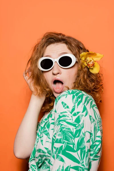 Mujer pelirroja joven conmocionada con flores de orquídea y gafas de sol posando en blusa con patrón floral y de pie sobre fondo naranja, verano casual y concepto de moda, Cultura Juvenil - foto de stock