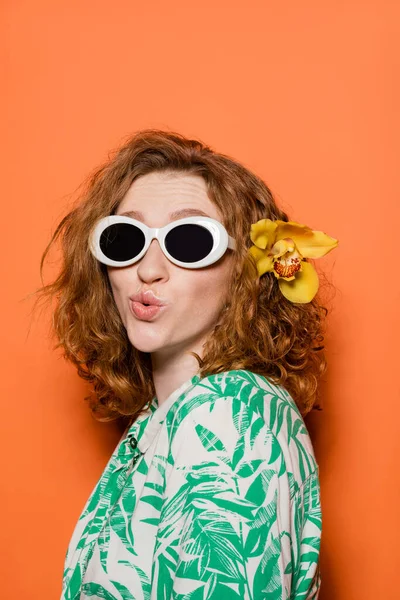 Mujer joven con pelo rojo y flor de orquídea con gafas de sol y blusa con patrón floral mientras hace pucheros labios y de pie sobre fondo naranja, verano casual y concepto de moda, cultura juvenil - foto de stock