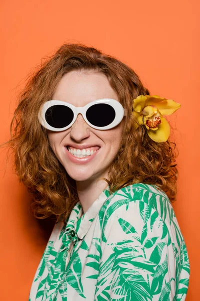 Positive Frau mit Orchideenblüte im roten Haar mit Sonnenbrille und Bluse mit floralem Muster, posierend und stehend auf orangefarbenem Hintergrund, sommerliches Freizeit- und Modekonzept, Jugendkultur — Stockfoto