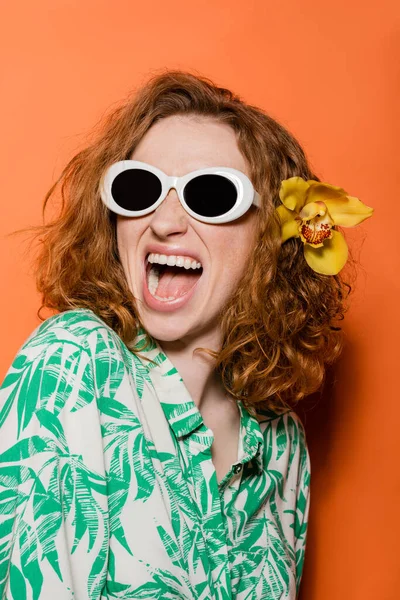 Excited redhead woman with natural makeup and orchid flower wearing sunglasses and blouse with floral print while standing on orange background, summer casual and fashion concept, Youth Culture — Stock Photo